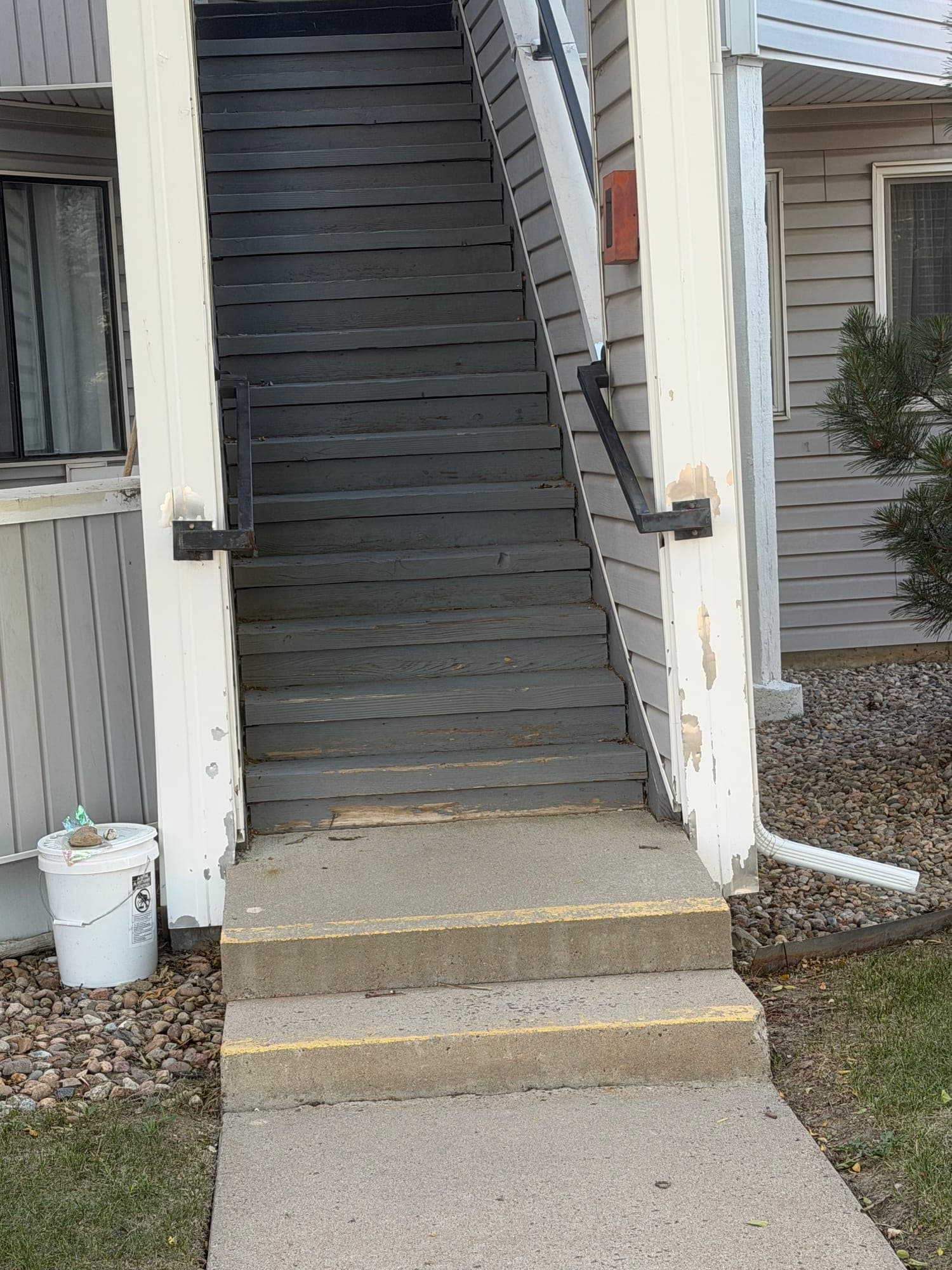 Stair base and downspout discharge — IMG_1048: June 2026 condition photo showing exterior stair base conditions beside a downspout and concrete walkway.