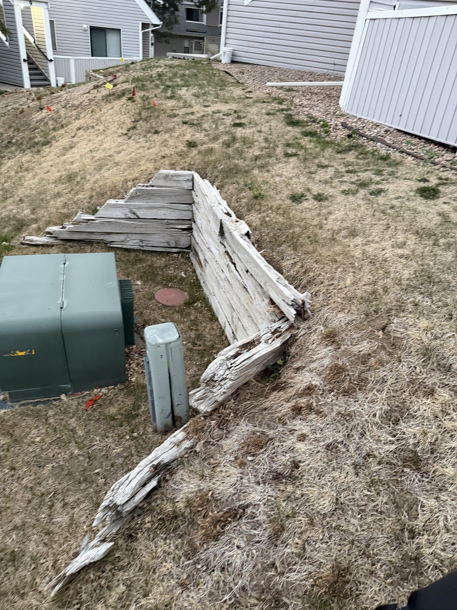 Failed retaining wall by utility box — IMG_1071: Collapsed timber retaining wall on a slope beside a green utility pedestal.