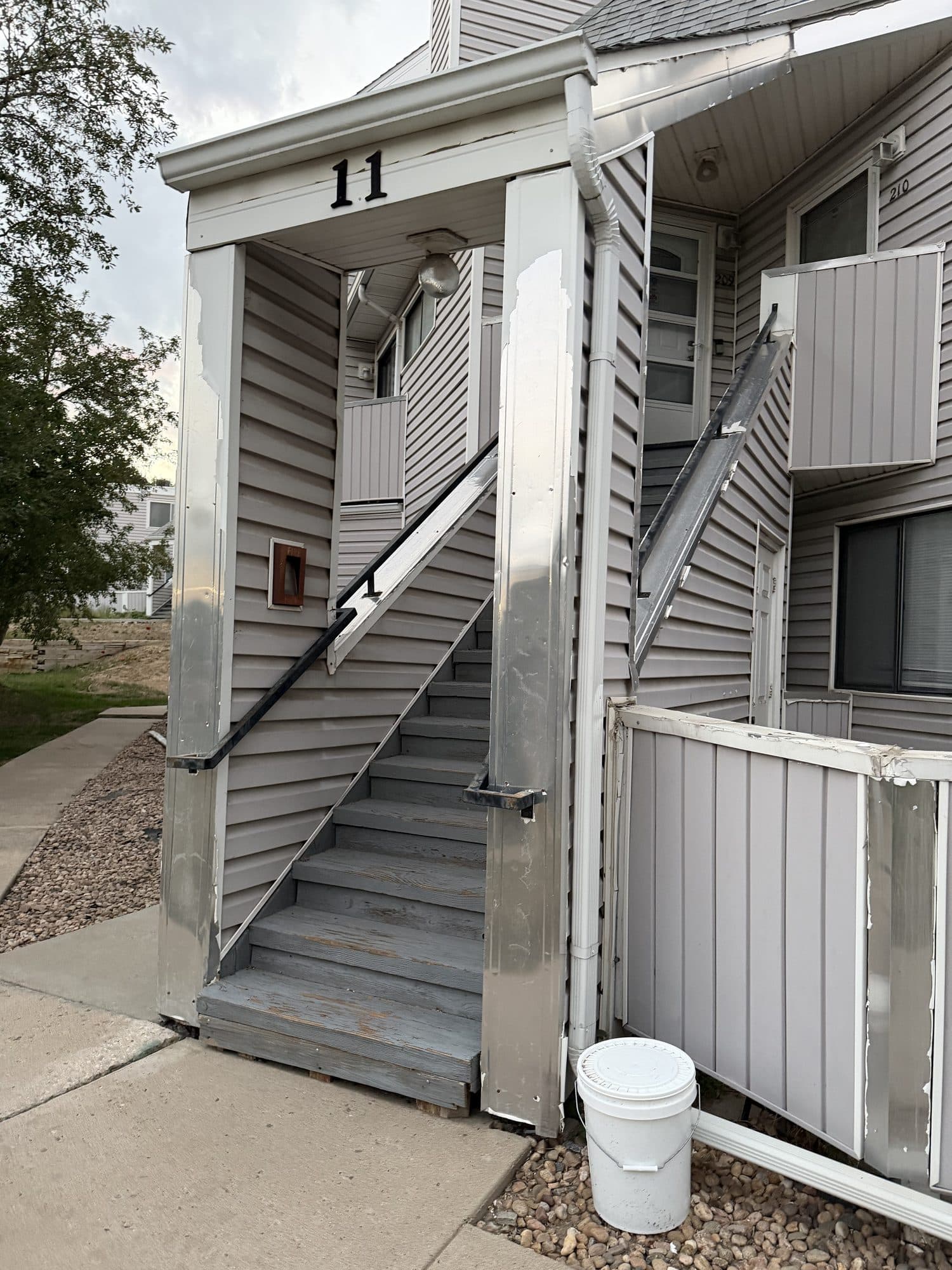 Failing clad stairway at unit 11 — IMG_1074: Exterior stairway at unit 11 with weathered treads, peeling paint, and bent aluminum cladding on posts and guards.