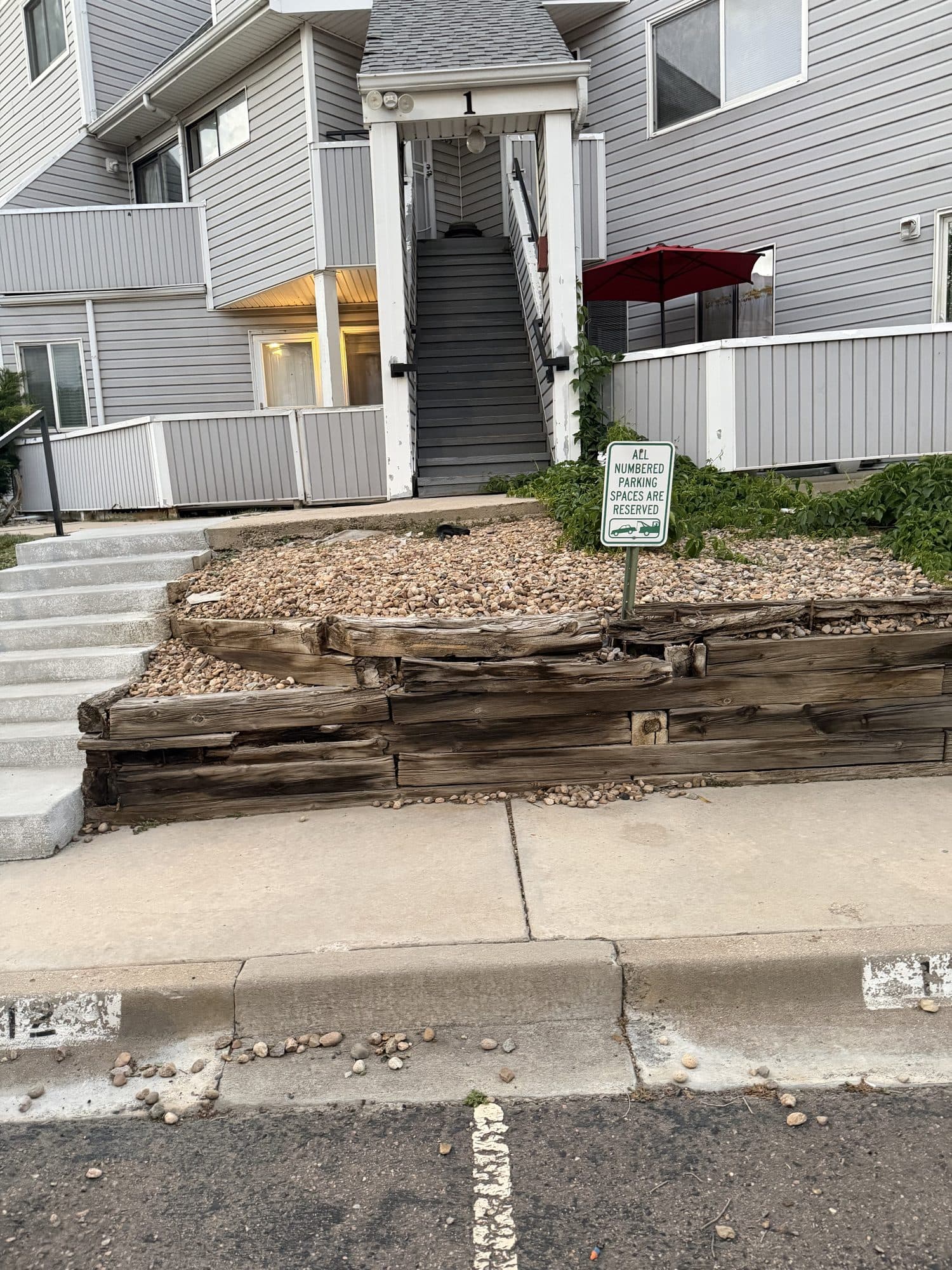 Collapsing timber retaining wall at unit 1 — IMG_1078: Severely deteriorated timber retaining wall at unit 1, with rotted timbers and gravel spilling onto the sidewalk and street.