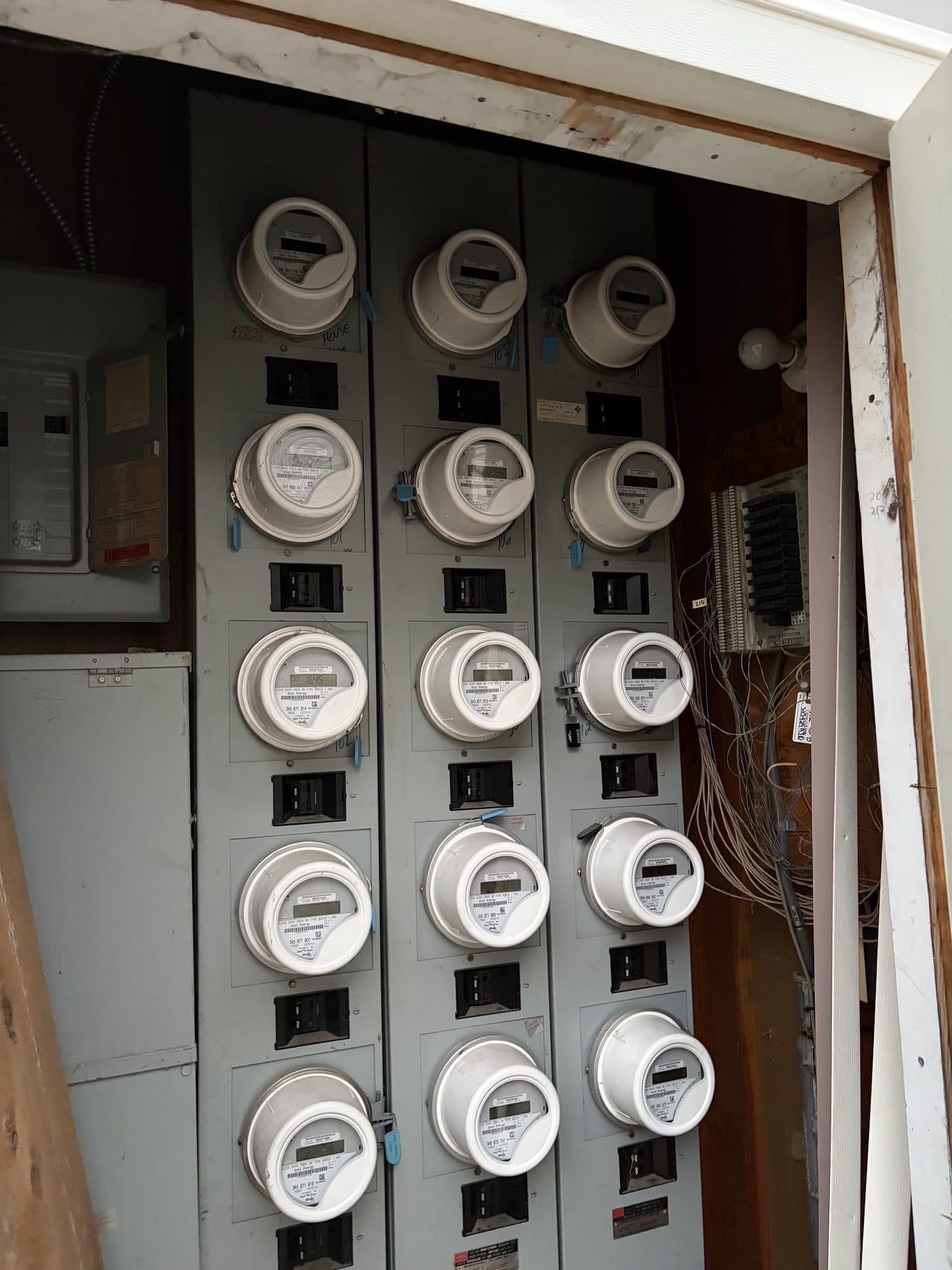 Communal meter stack with exposed wiring — IMG_6914: The communal meter stack (about 15 meters) with loose, exposed wiring and a displaced, hanging enclosure cover. This is common electrical service equipment; exposed conductors are a safety hazard.